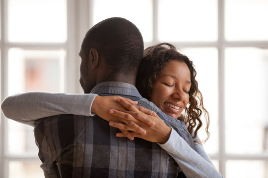 Attractive loving African American girlfriend embrace boyfriend, standing at home with closed eyes, affectionate couple in love romantic relationship, wife supporting husband rear view, gratefulness