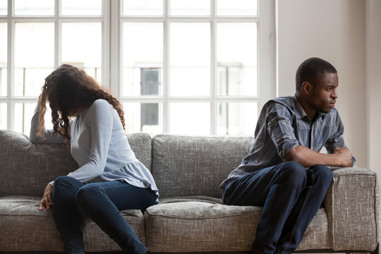 African American couple sitting separately on couch in living room after quarrel, upset woman hiding face in hair, frustrated man looking in distance, no desire to talk, relationship problem