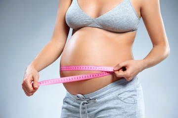 Close up of pregnant woman with measuring tape measures her belly on grey background. Pregnancy, maternity, preparation and expectation concept
