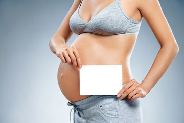 Close up of pregnant woman holding greeting card on grey background. Pregnancy, maternity, preparation and expectation concept