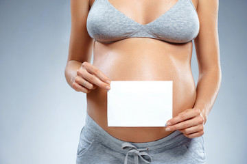 Close up of pregnant woman holding greeting card on grey background. Pregnancy, maternity, preparation and expectation concept