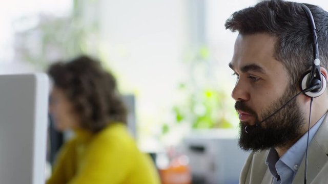 Tilt down of young bearded businessman working in call center: he using computer and headphones while talking with customer via phone call in the office