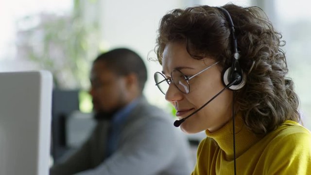 Close-up of cheerful Caucasian businesswoman in headphones using computer and talking with client while working in call center; African man working in background