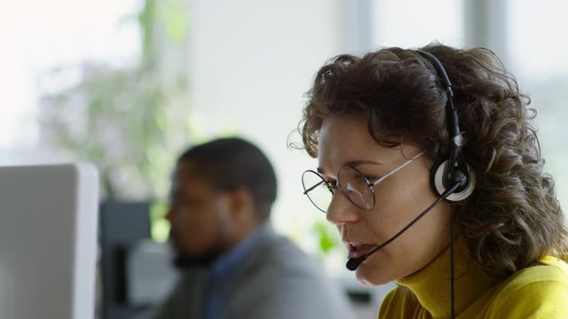 Tilt down of young Caucasian businesswoman in headphones using computer and talking with customer while working in call center; African man working in background