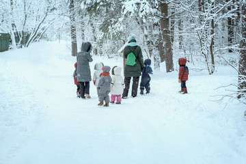 Several of unrecognizable little kids with teachers of kindergarten, back to us, walk holding hands in snowy park, winter leisure, active lifestyles