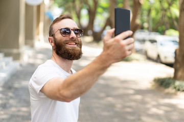 Joyful hipster guy taking selfie. Young man with bushy beard, wearing summer clothes and smiling at...