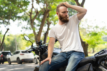Serious biker sitting on broken motorbike and looking to be helped. Bearded young motorcyclist holding head. Roadside assistance concept