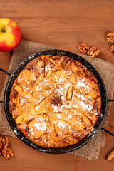 A closeup photo of an apple pie, shot from the top on a dark rustic wooden background with an apple, walnuts, anise, and a place for text