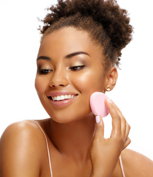Young girl applying foundation on her face using makeup sponge. Photo of smiling african girl on white background