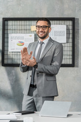 handsome happy businessman applauding and smiling at camera while standing in office