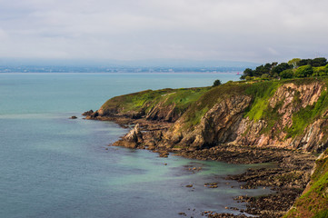 Fototapeta premium Irish coast landscape with rugged cliffs covered in green grass on a cloudy summer day. Howth Cliff Walk, County Dublin, Ireland.