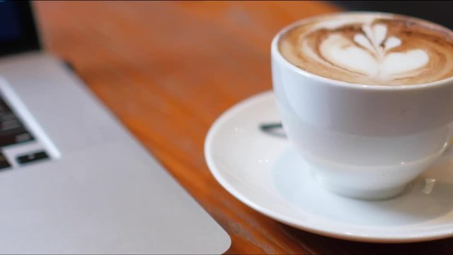 A cup of cappuccino coffee with laptop on table. Royalty high quality free stock footage of capuccino coffee with laptop for working in a coffee shop. Beautiful workspace with retro and vintage style