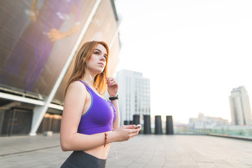 Street portrait of an attractive athletic girl with a smartphone in his hands listens to music in the headphones while jogging, looking away, standing for rest. Copyspace Sports concept.