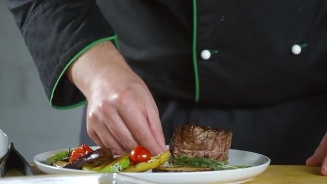 Close up shot of hands of unrecognizable restaurant chef plating fillet mignon. He is putting sauce dish and fresh thyme on plate with beef steak and vegetables