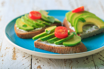 Avocado sandwich on dark rye toast bread made with fresh sliced avocado, cream cheese, seeds and cherry on white wooden background