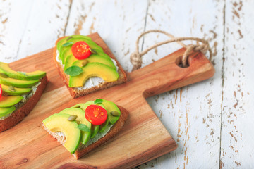 Avocado sandwich on dark rye toast bread made with fresh sliced avocado, cream cheese, seeds and cherry on white wooden background