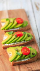 Avocado sandwich on dark rye toast bread made with fresh sliced avocado, cream cheese, seeds and cherry on white wooden background