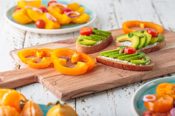 Avocado sandwich on dark rye toast bread made with fresh sliced avocado, cream cheese, seeds and cherry on white wooden background