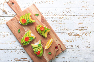 Avocado sandwich on dark rye toast bread made with fresh sliced avocado, cream cheese, seeds and cherry on white wooden background