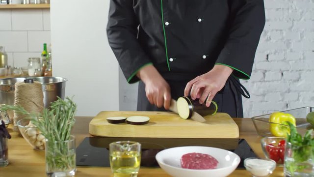 Mid-section tracking shot of unrecognizable restaurant chef cutting eggplant on wooden board in kitchen. Fresh vegetables, herbs and raw beef steak are on table