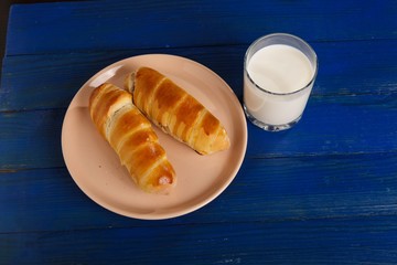 Sausage baked in the dough on a plate and a glass of milk on the table.