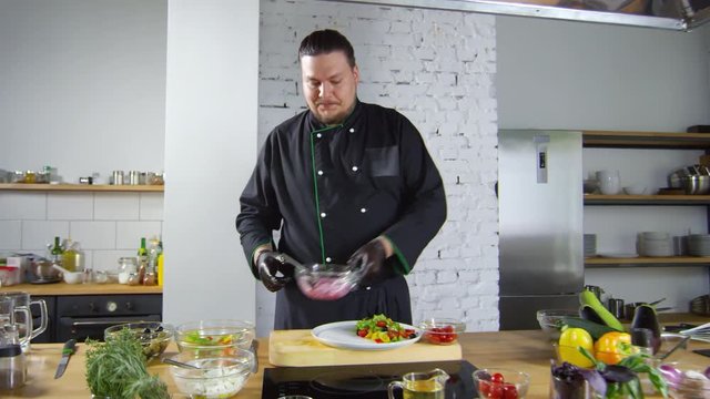 Tracking shot of male restaurant chef in uniform talking to camera and adding slices of red onion to salad on plate. Fresh vegetables and herbs are on table