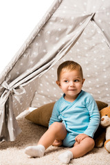 cute toddler boy smiling and sitting in baby wigwam with pillow and teddy bear isolated on white © LIGHTFIELD STUDIOS