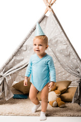 toddler boy in party hat standing in front of wigwam with pillows and teddy bear isolated on white © LIGHTFIELD STUDIOS