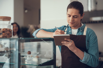 Young dark haired barista putting his elbow on the food glass case and looking attentive while writing in his notebook