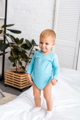 smiling baby in blue bodysuit standing on bed in nursery room