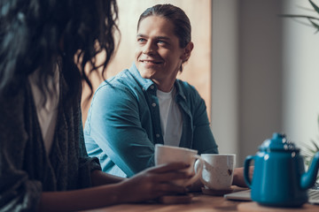 Positive handsome young man sitting close to his girlfriend and smiling while looking at her in the cafe