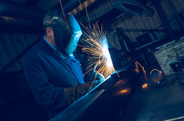 A vibrant action shot of a skilled working metal welder in action, welding metal. Photographed with a slow shutter speed and spark trails. Orange and teal. - obrazy, fototapety, plakaty