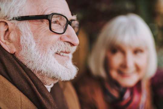 Close Up Side View Portrait Of Senior Man In Glasses Looking Away And Smiling. Old Lady On Blurred Background