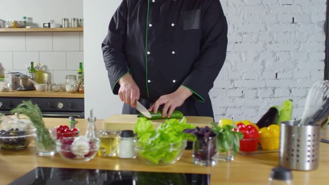 Tracking shot with tilt down of male professional cook in uniform cutting fresh cucumber while preparing salad in kitchen