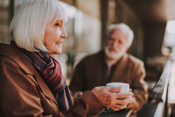 Side view portrait of joyful woman holding cup of coffee while looking away and smiling. Bearded gentleman on blurred background