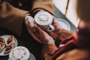 Cozy atmosphere. Close up of male and female hands holding cup of hot drink with foam. Pensioners...