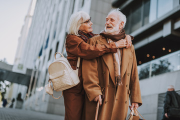 True love. Portrait of stylish lady with backpack hugging husband from behind while he looking at her and smiling
