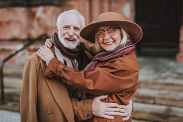Waist up portrait of stylish bearded man embracing happy wife. They looking at camera and smiling