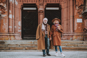 Fototapeta na wymiar Full length portrait of stylish bearded man and his wife standing near old building. Lady in hat holding cup of coffee