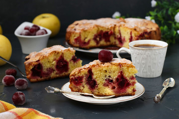 Berry pie with cherries is located on a plate on a dark background. In the foreground is a piece of cake and a cup of coffee