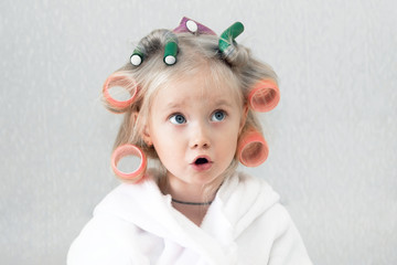 Girl with curlers hairpins in her hair. A little girl in a white coat spun pink curlers into her...