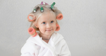 Girl with curlers hairpins in her hair. A little girl in a white coat spun pink curlers into her...