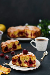 Berry pie with cherries is located on a plate on a dark background. In the foreground is a piece of cake and a cup of coffee