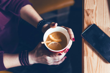 Top view. Student woman holds with two hands a hot cup of tea with sea buckthorn Hippophae and a slice of orange. Workplace, laptop, mobile phone, smartphone. coffee shop, warm color toning