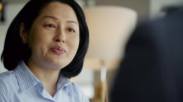 Close up face of friendly middle-aged Asian woman talking to unrecognizable colleague seen from their back, tracking left