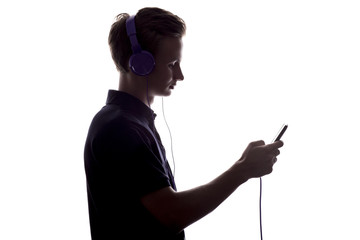 silhouette of a young man listening music in headphones, guy flips through a playlist on smartphone display on a white isolated background