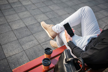 Obraz premium Cut view of man in jeans sitting on bench outside and hold black phone. There re two cups and leather bag.
