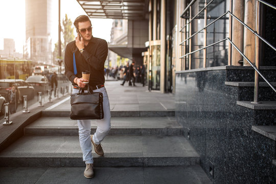 Young businessman walks on steps. He holds phone close to ear and care leather bag. Guy is serious. He wears sunglasses.