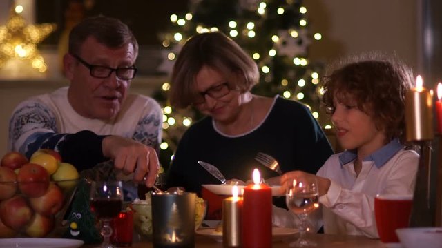 Happy family celebrating Christmas by candlelight against the background of New Year's lights. Grandpa puts salad in his grandson's plate. Vegetarian dinner