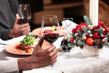 The hands of two African Americans holding glasses of red wine in the restaurant. Near hand in focus. The far arm is out of focus. Concept of rest. Christmas accessories on the table.